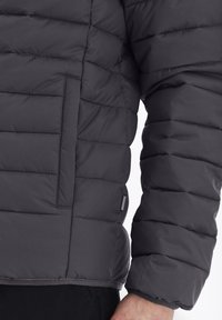 Grå puffjacka med quiltad textur, dragkedjeförsedd framficka och elastiska ärmslut. Tillverkad av lätt, isolerat material för värme.