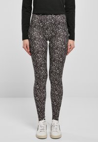 Leggings neri con motivi astratti bianchi, texture liscia, forma aderente, abbinati a sneakers bianche su uno sfondo neutro.