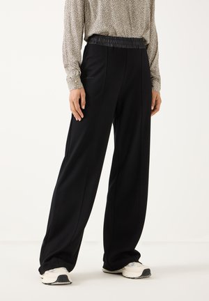 Personne portant un pantalon noir à jambes larges avec une ceinture élastique, associée à un chemisier clair à motifs et des baskets blanches sur un fond uni.