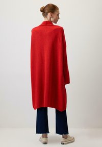 Červený oversize kardigán vyrobený z mäkkej, textúrovanej tkaniny. Má vysoký golier a bočné rozparky. Kombinovaný s tmavými džínsami a pohodlnými topánkami.