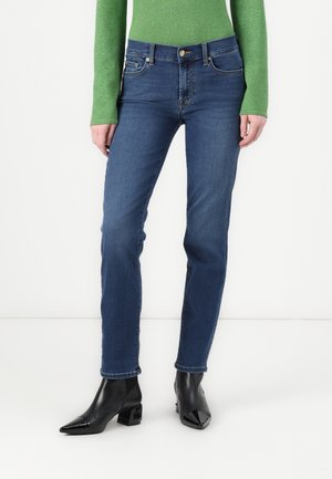 Femme portant un jean bleu droit, un haut vert à manches longues et des bottines noires à bout pointu sur fond blanc.