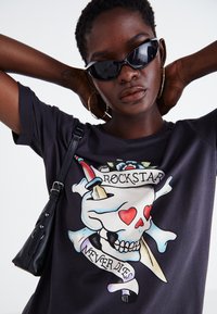 Juoda marškinėliai su spalvinga kaukolės grafika, kuri papildyta širdelėmis, tekstu "ROCKSTAR" ir "NEVER DIES", derinami su stilingais akinių nuo saulės ir maža kuprine.