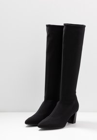 Bottes noires montantes en tissu doux, avec un bout pointu et un talon empilé. Texture lisse avec des détails de couture subtils.
