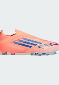 Botín de fútbol de color rosa brillante con parte superior texturizada, diseño aerodinámico, franjas azules como acento y tacos transparentes para una mejor tracción en el césped.