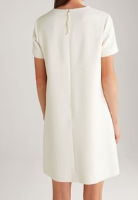 Kurzärmeliges, knielanges Kleid aus cremefarbenem Stoff mit glatter Textur. Verfügt über eine Rückennaht und einen Schlaufenknopfverschluss am Hals.