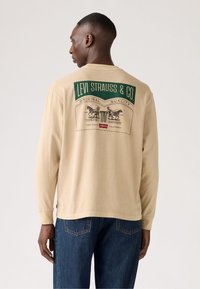 Beige lange mouwen T-shirt met een grote gedrukt Levi Strauss & Co. graphic op de achterkant met donkergroene en zwarte accenten.
