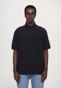 Marineblå kortermet polo med strukturert mønster, tre knappelukking og mørk krage. Kombinert med lyseblå denimjeans.