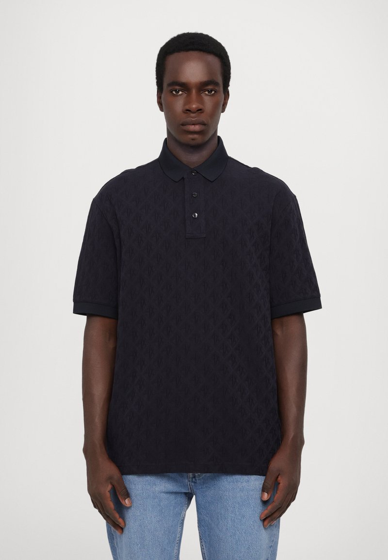 Marineblå kortermet polo med strukturert mønster, tre knappelukking og mørk krage. Kombinert med lyseblå denimjeans.