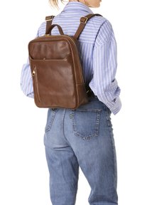 Sac à dos en cuir marron de forme rectangulaire, avec fermeture éclair et poche avant. Texture lisse, bretelles réglables et détails de couture subtils.