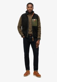 Sort fleece vest over en tekstureret brun sweater med en oliven grøn lomme. Mørke jeans og brune sko, stående mod en ensfarvet baggrund.