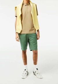 Hnědé polo tričko, zelené kraťasy, bílé tenisky s černými akcenty a žlutý svetr přehozený přes ramena. Uvolněný letní outfit.
