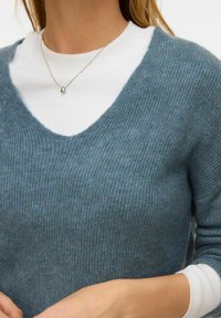 V veida kakla trikotāžas džemperis no mīkstas ribveida auduma tumši tējas zaļā krāsā, uzvilkts pāri baltai ar apaļu kakla izgriezumu garroku krekliņam. Sudraba kaklarotas detaļa.