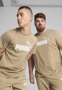Two men wear beige cotton t-shirts with large white "PUMA" branding. The shirts are short-sleeved with a crew neck design and relaxed fit.