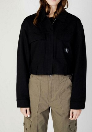 Žena nosí černou krátkou bundu s našitými kapsami a béžové vysoké kalhoty s bočními kapsami na knoflíky.