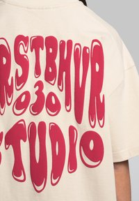 Béžové tričko s krátkým rukávem, na zadní straně s výrazným, vlnitým červeným textem "FRSTBHVR 030 STUDIO", který zdůrazňuje grafický design.