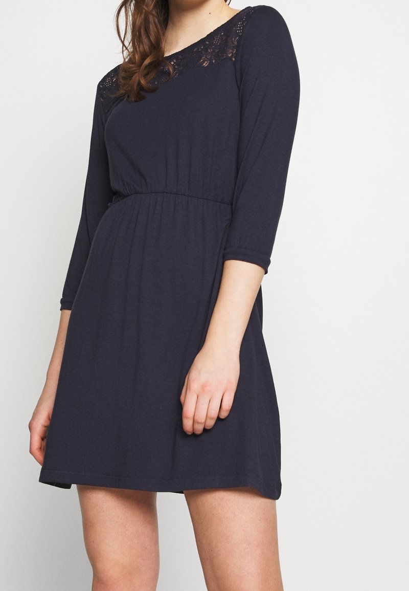 Femme portant une robe bleu marine foncé avec manches trois-quarts et un détail en dentelle au niveau du décolleté, debout devant un fond clair uni.
