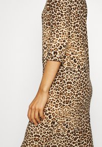 Le bras et le torse d'une femme portant une robe moulante à imprimé léopard avec des manches trois-quarts, sur un fond blanc uni.