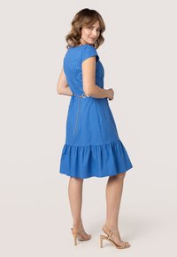 Blauw katoenen jurk met een aansluitende buste en een uitlopende gerimpelde zoom, met een kettingdetail op de rug en korte mouwen. Model gecombineerd met beige hakken.