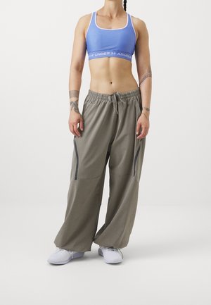 Vrouw draagt een blauwe sportbeha, losse grijze broek met trekkoord, witte sneakers en zichtbare tatoeages op haar armen, staand tegen een witte achtergrond.