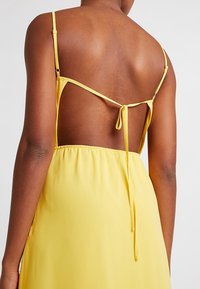 Robe jaune dos nu avec fines bretelles, taille froncée et détail de lien ajustable à l'arrière. Tissu lisse avec une finition brillante.