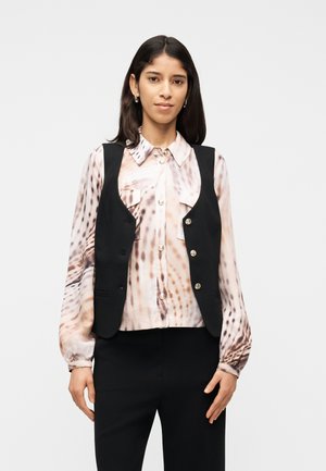 Nő, aki mintás hosszú ujjú blúzt, fekete gombos mellényt és fekete nadrágot visel, egy egyszerű világos háttér előtt áll.