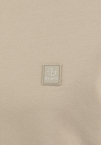 Tejido beige texturizado con un patrón diagonal; presenta un emblema cuadrado plateado estampado con "KEY LARGO". Diseño simple y superficie suave.