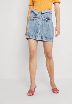 Kvinde iført en blå denim nederdel med knapper og en foldet linning, en orange kortærmet top og lyserøde hælede sandaler.