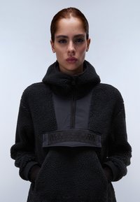 Sort fleece-pullover med høj krave, der har et mat stofpanel og et fremtrædende sort logo-patch foran. Tekstureret, hyggeligt materiale.