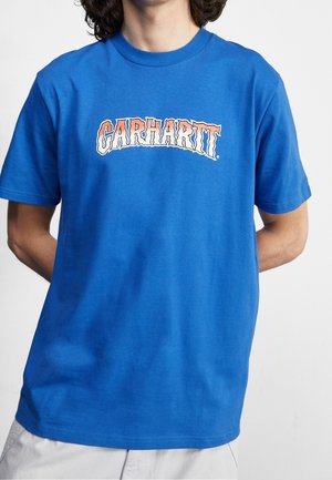 Person trägt ein blaues kurzärmeliges T-Shirt mit dem Schriftzug „CARHARTT“ in rotem und weißem Flammenstil auf der Brust.