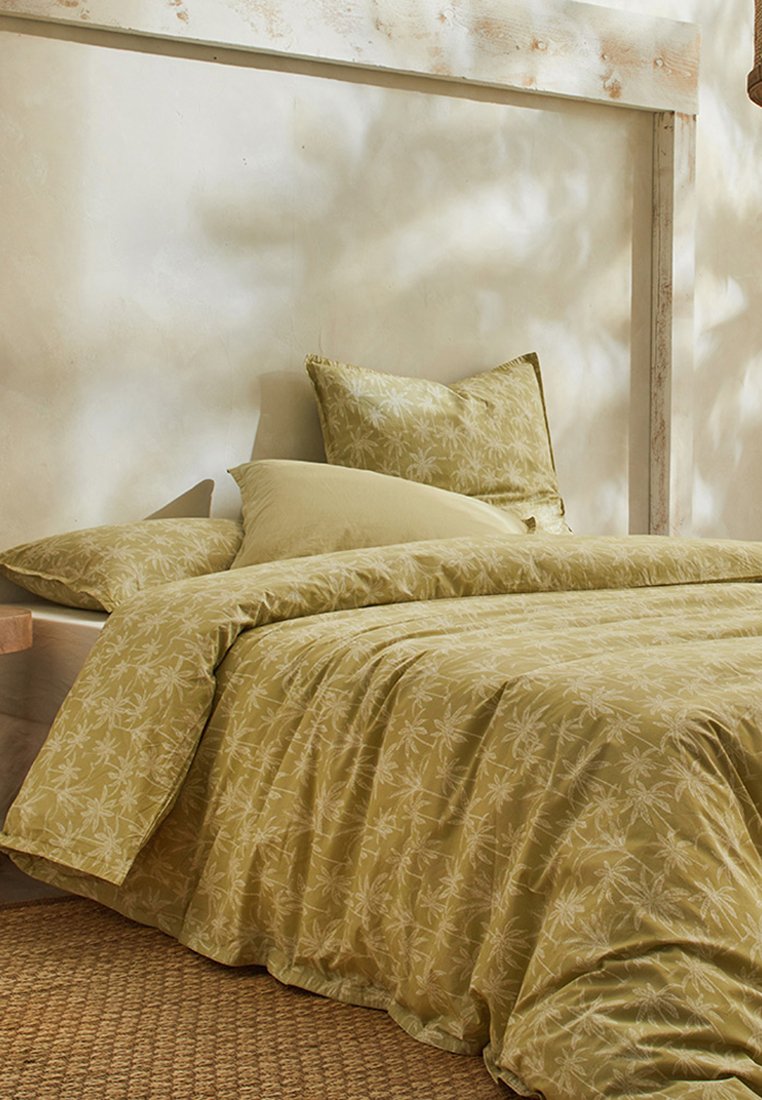 Housse de couette et coussins vert olive ornés d'un subtil motif de feuilles sur un tissu doux, disposés soigneusement sur un cadre de lit en bois avec un tapis texturé.