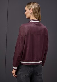Veste bomber bordeaux en tissu doux, avec un col et des poignets côtelés rayés blancs et noirs, présentant un détail de couture sur l'épaule arrière.