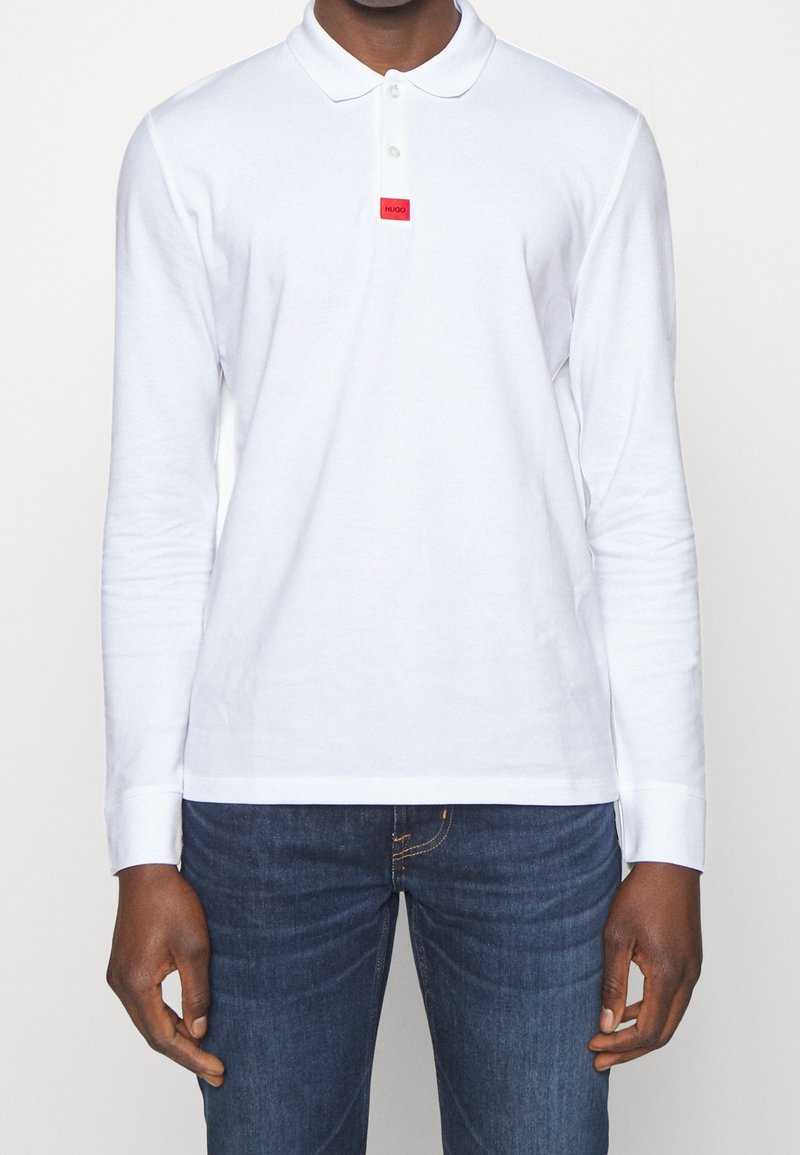 Weißes Langarm-Polohemd mit klassischem Kragen, ausgestattet mit einem roten Logopatch, aus einem glatten Stoff hergestellt und kombiniert mit dunklen Denim-Jeans.