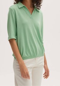 Světlonzelený polo top s krátkým rukávem a límečkem, se elastickým pasem, s hladkou texturou a ohrnutými rukávy. Nosený s bílými kalhotami.