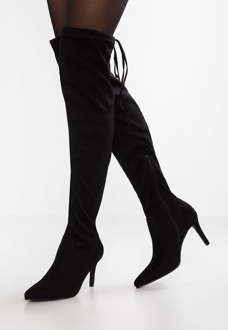 Žena v černých semišových kozačkách po kolena se špičatou špičkou a úzkým podpatkem, doplněná černými průhlednými punčochami.