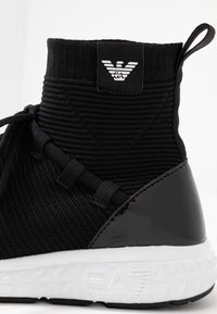 Sneaker alte nere con tomaia in maglia testurizzata, dettagli neri lucidi e una suola in gomma bianca con un motivo geometrico.