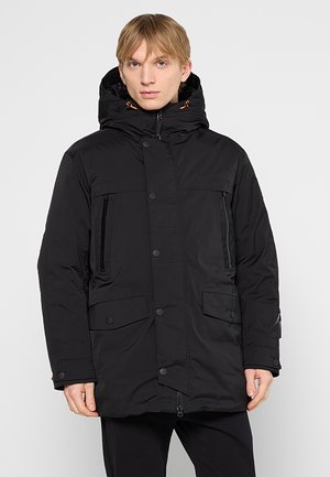 Junger Mann trägt eine schwarze winterliche Kapuzenjacke mit Reißverschluss und Knöpfen, steht vor einem schlichten hellgrauen Hintergrund.