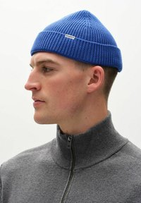 Blå ribbad stickad mössa med spetsig topp och logotags, bärs tillsammans med en grå zip-up tröja. Fokusera på textur och färgernas kontrast.