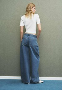 Persona bionda che indossa una maglietta bianca a maniche corte, jeans blu a vita alta larghi e scarpe bianche a punta, in piedi contro un muro grigio.