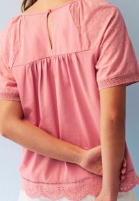 Blouse rose clair avec des manches courtes brodées, un dos à ouverture en goutte d'eau avec fermeture à bouton, et un ourlet en dentelle à volants.