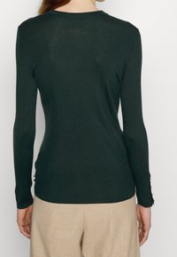 Kobieta ubrana w dopasowaną, ciemnozieloną bluzkę z długim rękawem i beżowe spodnie, pokazana z tyłu na tle jednolitego białego tła.