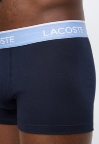 Κοντινό πλάνο ενός ατόμου που φοράει σκούρο μπλε μποξέρ με ανοικτού μπλε μέση όπου αναγράφεται το "Lacoste".