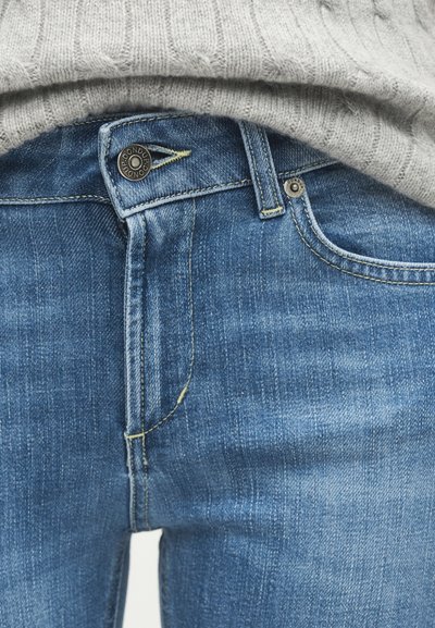 Μεσαίου μπλε τζιν παντελόνι με κούμπωμα κουμπιού και θηλιές για ζώνη, σε συνδυασμό με ανοιχτό γκρι ριμπ πλεκτό πουλόβερ που είναι μπακαλισμένο μέσα.