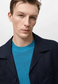 Marineblaues Jacket mit zwei Vorderaschen über einem gerippten, helltürkisfarbenen Pullover. Glatte Stofftextur mit klassischem Kragen Design.