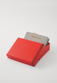 Portafoglio in pelle grigia con una finitura textured e lettering dorato "VALENTINO", parzialmente visibile in una scatola rossa. La scatola ha una superficie liscia.