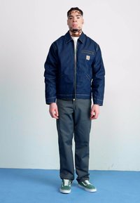 Marineblå denimjakke med glidelås og brystlomme, kombinert med mørkegrå bukser og grønne sneakers. Plassert mot en enkel bakgrunn.