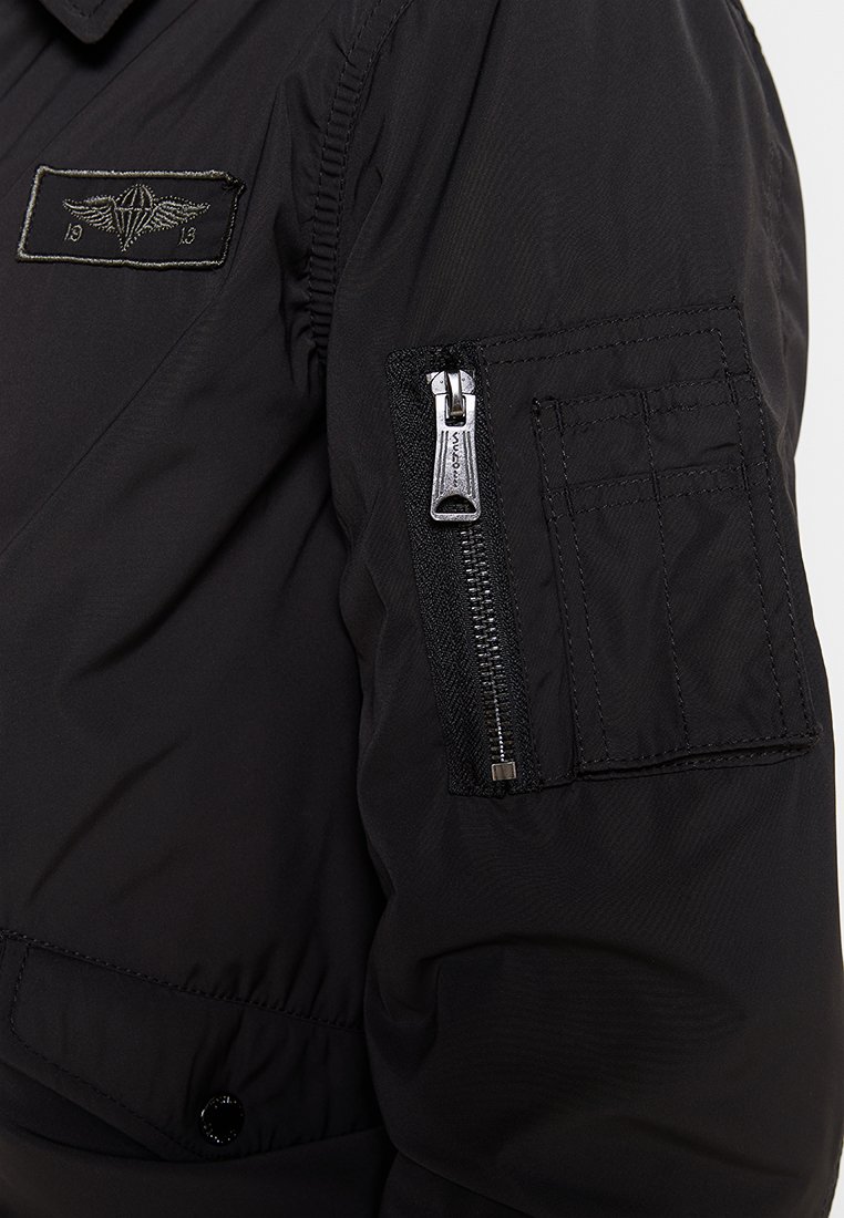 Veste bomber noire avec une poche zippée sur la manche gauche, décorée d'un patch en tissu avec des ailes et les chiffres "19 13". Texture lisse et durable.