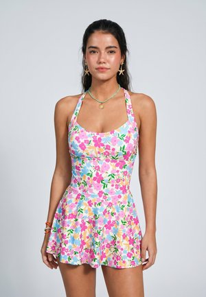 Frau steht und blickt nach vorne, trägt ein ärmelloses Kleid mit auffälligem pinken, gelben, grünen und blauen Blumenmuster, schlichter Hintergrund.