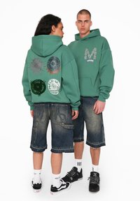 Grøn bomulds hoodie med grafiske tryk på ryggen, parret med mørke denim shorts. Outfittet inkluderer sneakers og hvide sokker.