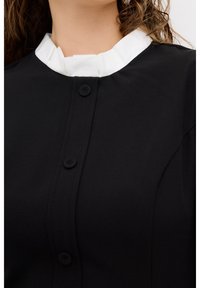 Czarna bluzka z materiału z falbaniastym białym kołnierzykiem, z zapięciem na guziki z przodu i gładką strukturą. Zbliżenie na dekolt i guziki.