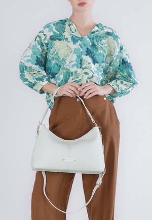 Persoon draagt een groene blouse met bloemenpatroon en bruine broek, en houdt een witte leren handtas met twee hengsels vast voor een effen achtergrond.